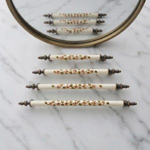 May include: A set of seven off-white ceramic drawer pulls arranged in a vertical formation. Each pull features a hand-painted floral pattern with small pink flowers and green leaves, finished with dark metallic finials at both ends.