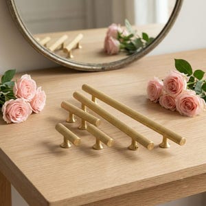 May include: A collection of four gold-toned metal cabinet pulls and knobs with a textured, knurled surface. The hardware pieces vary in length, featuring cylindrical bars supported by small, circular pedestals, arranged on a light wood surface.