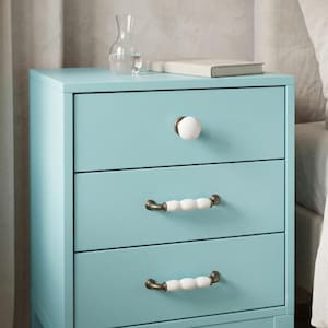 May include: A light blue bedside table with three drawers. The top drawer features a single round white knob, while the two lower drawers each have a horizontal handle with a white, segmented, bead-like design and metallic ends.