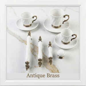May include: A collection of white ceramic cabinet hardware with antique brass accents, including three varying lengths of drawer pulls and one round knob, displayed alongside three white ceramic teacups and saucers with brass bases.
