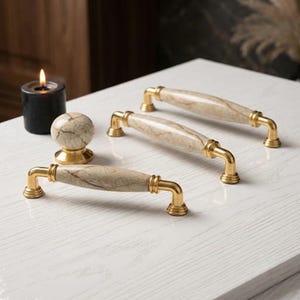 May include: Two elongated cabinet pulls and one round cabinet knob featuring a beige marble-patterned center with gold-toned metal ends and mounting bases. The hardware is arranged on a white surface next to a small black candle with a lit flame.