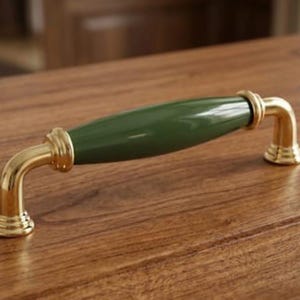 May include: A cabinet pull handle featuring a central olive green ceramic barrel section with a glossy finish, flanked by polished brass metal ends that curve downward into circular mounting bases, creating a smooth and rounded hardware piece.