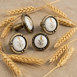 May include: Four round cabinet knobs featuring a white ceramic center with a painted golden wheat stalk design. Each knob is encased in an antique brass-toned metal frame with decorative scrollwork details along the circular outer edge.
