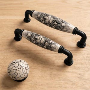 Può includere: Due maniglie per armadietto arcuate e un pomello rotondo. Ogni pezzo presenta un centro in ceramica color crema con un motivo floreale nero e finiture in metallo nero alle estremità. Le maniglie hanno un design curvo con basi di montaggio nere.