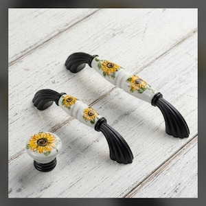 May include: Two cabinet pulls and one round cabinet knob featuring a white ceramic center with yellow sunflower patterns and green leaves. Each piece has black, ribbed metal ends with a curved, fluted design for mounting to furniture.
