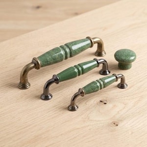 May include: Three cabinet pulls and one round cabinet knob. The pulls feature green ceramic centers with a marbled texture and metallic ends. The largest pull has an antique brass finish, while the two smaller pulls have a dark metallic finish.