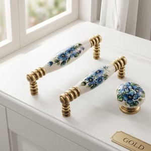 May include: Two ceramic drawer pulls and one matching round knob, each featuring a white base with hand-painted blue flowers and green leaves, accented by ribbed antique gold metal hardware at the ends and base.
