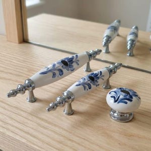 Può includere: Due maniglie per cassetti in ceramica bianca e un pomello rotondo in ceramica, ciascuno caratterizzato da un motivo floreale blu dipinto a mano e da finiture in metallo color argento lucido. Le maniglie allungate hanno terminali decorativi.