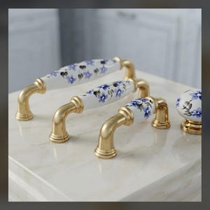 May include: Three cabinet pulls and one round cabinet knob featuring white ceramic centers with hand-painted blue floral patterns and green leaves. Each piece has a brushed gold metal finish on the mounting hardware and base components.