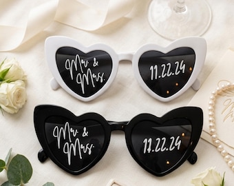 May include: Two pairs of heart-shaped sunglasses, one white and one black, featuring white script text on the lenses. The left lenses display "Mr. & Mrs." and the right lenses display the date "11.22.24" in a clean, cursive font style.