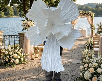 May include: A large, white, sculptural garment featuring an oversized, pleated, flower-like structure that covers the upper body and extends outward, paired with a matching pleated skirt and dark trousers, creating a dramatic, voluminous silhouette.