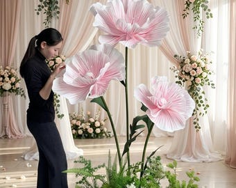 May include: Three oversized, artificial pink flowers with layered, pleated petals mounted on tall, slender green stems. The flowers are arranged in a cluster, emerging from a base of lush green foliage and ferns, creating a decorative floral display.