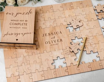 May include: A light-colored wooden guest book puzzle with interlocking pieces. A vertical sign reads, "our puzzle WOULD NOT BE COMPLETE WITHOUT YOU please sign a piece." The center features "JESSICA and OLIVER 08.09.2027." A gold pen rests on the puzzle.