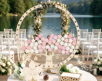 May include: A circular wooden guest book stand featuring a tree design with pink and white flower-shaped wooden pieces. The base includes the text 'Simon & Voice 09.09.2023'. A small sign reads 'Please sign your name and give best wishes for Mr & Mrs'.
