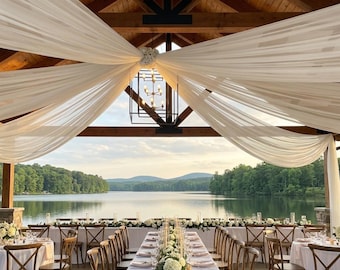 May include: An outdoor event space featuring white fabric draped from a wooden ceiling, a black metal lantern chandelier, and long tables covered in white tablecloths with floral centerpieces, overlooking a calm lake and distant mountains.