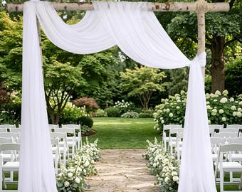 May include: A wedding arch constructed from two vertical birch wood poles and one horizontal crossbar, draped with long, flowing white sheer fabric. The fabric is gathered at the top corners and hangs down to the ground on both sides.
