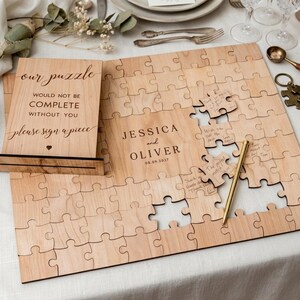 May include: A wooden puzzle guest book with the text "JESSICA and OLIVER 08.09.2027" engraved on it. The puzzle pieces are light brown. A sign reads "our puzzle would not be complete without you please sign a piece". A gold pen is included.