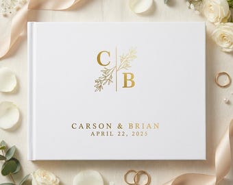 May include: A white hardcover guest book featuring gold foil lettering with the initials C and B separated by a vertical line and botanical sprigs. Below the initials, the text reads CARSON & BRIAN and the date APRIL 22, 2025.
