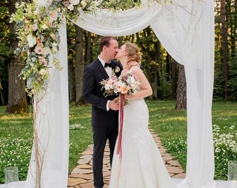 May include: A wedding arch draped in white fabric and decorated with a large floral arrangement of white, peach, and pink flowers with greenery. A person in a black suit and a person in a white lace wedding dress stand beneath the arch.