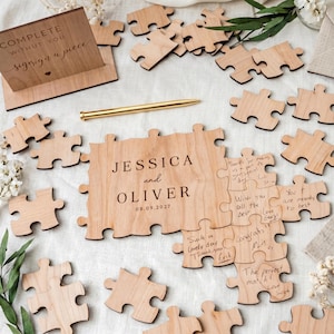 May include: A wooden puzzle guest book with the names "JESSICA and OLIVER" and the date "08.09.2027" engraved on it. The puzzle pieces are scattered around, with some signed. A gold pen and a sign that says "COMPLETE WITHOUT YOU signing a piece" are also present.