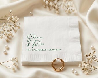 May include: A square white paper napkin featuring dark green printed text that reads Steven & Rose, THE CAMPBELLS | 08.08.2026. The napkin has a textured border and is accompanied by two gold hoop earrings and small white floral sprigs.