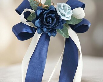 May include: A floral corsage featuring a large dark navy blue rose and a smaller light blue rose, accented with green leaves and gold sprigs. The arrangement is tied with long, flowing navy blue and white satin ribbons in a decorative bow.