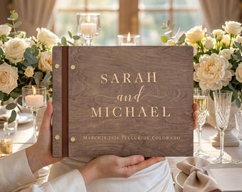 May include: A rectangular wooden photo album with a dark brown leather spine secured by gold metal rivets. The front cover features the gold-colored text "SARAH and MICHAEL" and "MARCH 28, 2026 TELLURIDE, COLORADO" engraved on the wood grain surface.