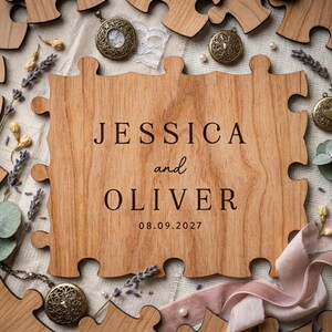 May include: A wooden puzzle piece engraved with "JESSICA and OLIVER" and the date "08.09.2027". The puzzle is surrounded by other wooden pieces, dried lavender, and vintage lockets.