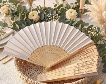 May include: A white folding hand fan with light wood ribs, featuring laser-engraved text reading Lee & Ofir 18/09/2025 and Clio & Mikey 19/09/2025 on the handle. The fan is displayed in a woven basket, showcasing a pleated design and intricate cutouts.