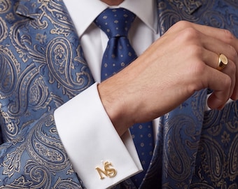 May include: A blue paisley blazer with a white dress shirt, a blue patterned necktie, and gold-toned metal cufflinks featuring the letters NS. A gold signet ring is worn on the finger, completing the formal attire with a polished appearance.