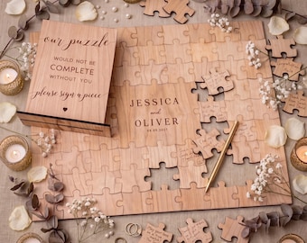 May include: A light wood guest book puzzle featuring the text "our puzzle WOULD NOT BE COMPLETE WITHOUT YOU please sign a piece" and "JESSICA and OLIVER 08.09.2021". Includes a gold pen, loose puzzle pieces, and several small glass candle holders.