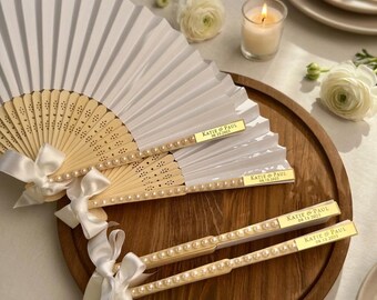 May include: A white folding hand fan with a light wood frame, featuring a gold rectangular plaque engraved with "Katie & Paul 08.18.2025". The handle is decorated with a row of white faux pearls and a white satin ribbon tied in a bow at the base.