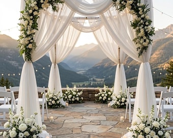 May include: A wedding arch constructed from four wooden posts draped with flowing white sheer fabric. The structure is adorned with large arrangements of white roses and greenery. White chairs and floor candles are placed on a stone patio.