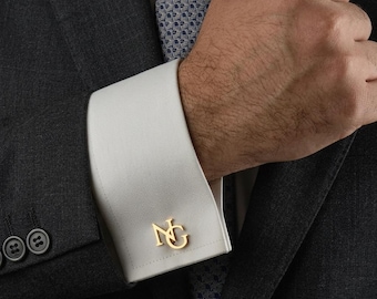 May include: A gold-toned metal cufflink featuring the interlocking letters NG, attached to a crisp white French cuff of a dress shirt. The shirt is worn under a dark grey textured suit jacket with a patterned tie visible at the collar.