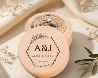 May include: A round light-colored wooden ring box with a lid featuring a black geometric frame, a leaf sprig, the initials A&J, the names ALICE & JADEN, and the date 04.04.2024. Inside the box, a silver ring with a clear stone sits on a jute insert.