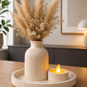 Könnte beinhalten: Eine cremefarbene Dekoration mit einer Vase mit vertikalen Rillen, einem Tablett und einem Kerzenhalter. Die Vase enthält einen Strauß getrockneter Pampasgras und kleine Blüten. Eine batteriebetriebene Kerze befindet sich auf dem Tablett und erzeugt eine warme Atmosphäre.