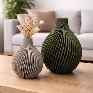 Könnte beinhalten: Zwei dekorative Vasen mit geripptem Design stehen auf einem Holztisch. Eine Vase ist hellgrau und enthält getrocknete Blumen. Die andere Vase ist dunkel olivgrün. Die Vasen stehen vor einem weißen Sofa.