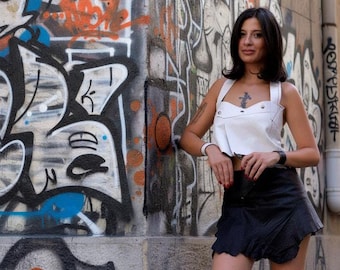 Può includere: Un top corto bianco senza maniche con corpetto strutturato e bottoni color argento, abbinato a una minigonna nera con finitura testurizzata e orlo smerlato, indossato con un cinturino nero e un braccialetto bianco.