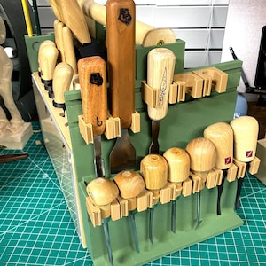 May include: A sage green 3D-printed tool organizer rack holding various wood carving chisels and gouges with light-colored wooden handles. The rack features multiple slots and clips to secure the metal-tipped tools in an upright position.