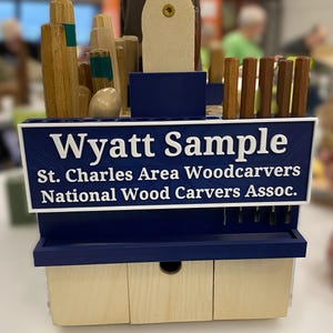 Puede incluir: Un expositor de madera con varias herramientas y muestras de tallado en madera. El expositor tiene un letrero azul marino que dice "Wyatt Sample" y "St. Charles Area Woodcarvers National Wood Carvers Assoc." El expositor está hecho de madera clara.