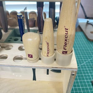 May include: A wooden tool organiser holding carving tools. Three Flexcut USA tools are visible, with wooden handles and the Flexcut logo. The organiser is made of light-coloured wood and has a handle.