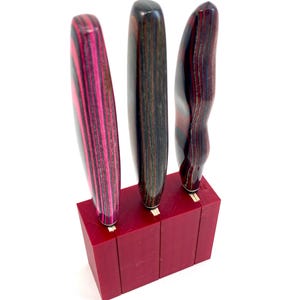 Peut inclure: Un bloc de couteaux rouge avec trois fentes, contenant trois manches de couteaux décoratifs. Les manches présentent un motif rayé dans les tons de rose, marron et noir. Le texte "Knives Not Included" est en bas.