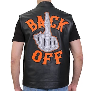 Può includere: Un gilet in pelle nera con una grande toppa ricamata sul retro. La toppa mostra una mano scheletrica grigia che fa il gesto del dito medio, circondata dalla scritta arancione brillante "BACK OFF" in un carattere audace e stilizzato.