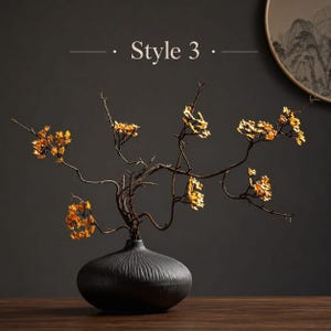 May include: A matte black ceramic vase with a ribbed texture and a wide, rounded base. The vase holds thin, dark brown branches adorned with small, vibrant yellow blossoms. The text "Style 3" is visible in the upper center of the image.