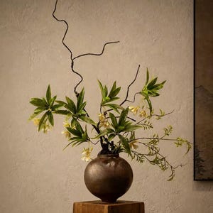 Peut inclure: Un vase sphérique de couleur bronze contenant des fleurs et des branches vertes et jaunes. Le vase est posé sur un socle carré en bois. Le texte "STYLE 1" est visible.