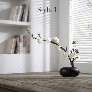 May include: A black, smooth, oval-shaped ceramic vase holds a single branch with white blossoms and green buds. The text "Style 1" is visible above the branch. The vase is positioned on a wooden surface, showcasing a simple, clean design.