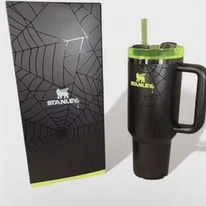 May include: Black Stanley travel mug with a green lid and handle, next to a matching box. The mug and box feature a spiderweb design and the Stanley logo. The mug includes a straw.