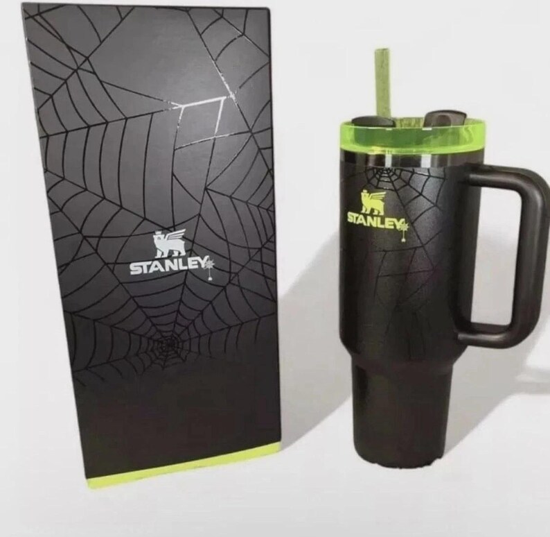 May include: Black Stanley travel mug with a green lid and handle, next to a matching box. The mug and box feature a spiderweb design and the Stanley logo. The mug includes a straw.