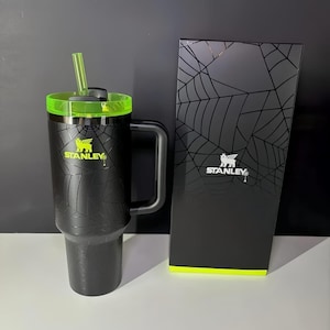 May include: Black Stanley tumbler with a neon green lid and straw, featuring a spiderweb design. The tumbler has a handle and the Stanley logo in neon green. A matching black box with a spiderweb design and the Stanley logo is next to the tumbler.
