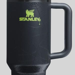 May include: Black Stanley tumbler with a green lid and straw. The tumbler features a spiderweb design and the Stanley logo in green. The handle is black. A Halloween-themed drinkware item.